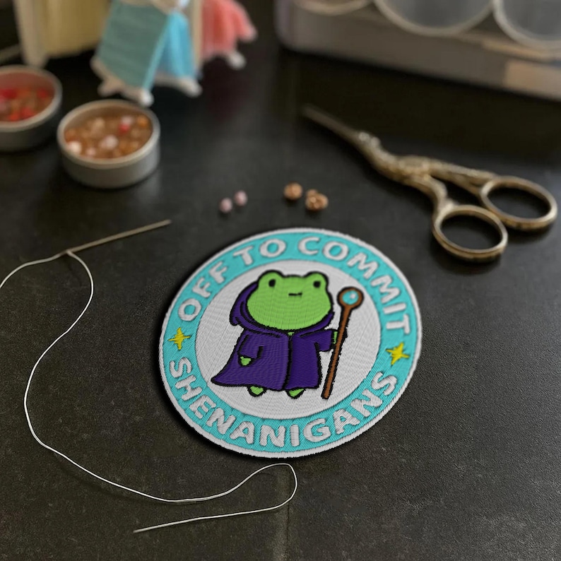 K&ouml;nnte beinhalten: Gestickter Aufn&auml;her mit einem Cartoon-Frosch in einem lila Gewand, der einen Stab h&auml;lt. Der Aufn&auml;her tr&auml;gt den Text "OFF TO COMMIT SHENANIGANS" in einem kreisf&ouml;rmigen Design mit t&uuml;rkisfarbenen und gelben Akzenten. Eine Nadel und Faden, eine Schere und kleine Beh&auml;lter befinden sich im Hintergrund.