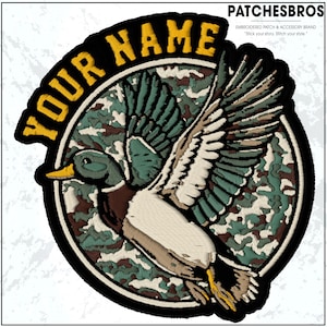 May include: Embroidered patch featuring a detailed mallard duck in flight, set against a camouflage background. The patch is circular, with the words "YOUR NAME" arched across the top in yellow. The brand name "PATCHESBROS" is also visible.