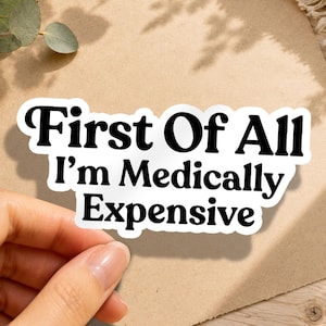 May include: A die-cut vinyl sticker with a white background and a black border. The sticker features the bold, black, serif-style text that reads: First Of All I'm Medically Expensive. The design is presented in a clean, high-contrast graphic format.