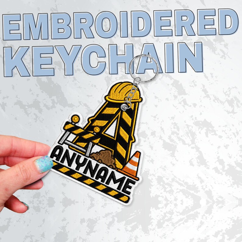 May include: An embroidered keychain featuring a construction-themed letter A with yellow and black hazard stripes, a yellow hard hat, a small orange traffic cone, and a pile of dirt, with the text ANYNAME printed in bold black capital letters.