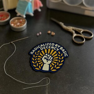 May include: A navy blue and yellow patch with the text "RADICALIZED BY BASIC DECENCY" and a raised fist design. The patch is surrounded by a gold laurel wreath and sunburst. Also visible are sewing supplies, including a needle, thread, and small scissors.
