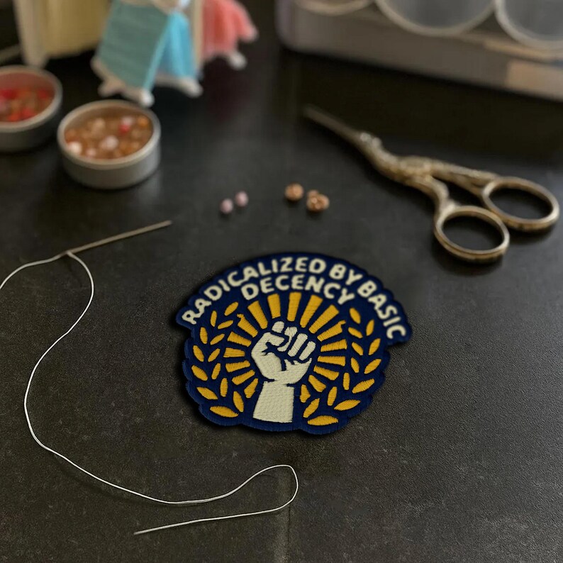 May include: A navy blue and yellow patch with the text "RADICALIZED BY BASIC DECENCY" and a raised fist design. The patch is surrounded by a gold laurel wreath and sunburst. Also visible are sewing supplies, including a needle, thread, and small scissors.