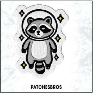 May include: Embroidered patch featuring a cartoon raccoon in a spacesuit, surrounded by yellow stars. The raccoon is gray, white, and black, with a detailed design. The text "PATCHESBROS" is at the bottom.