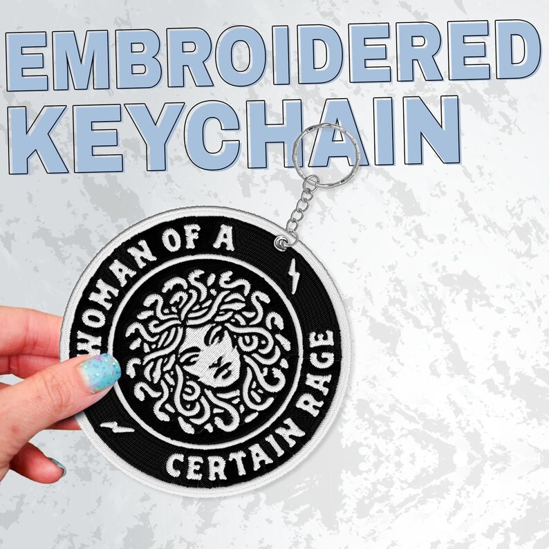 May include: Embroidered keychain with a black and white circular design. The design features a Medusa illustration and the text "WOMAN OF A CERTAIN RAGE". The keychain has a silver metal ring.