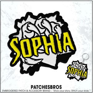 May include: Embroidered patch and keychain featuring the name "SOPHIA" in yellow, outlined in black, with a claw design. The patch and keychain are from the brand PATCHESBROS.
