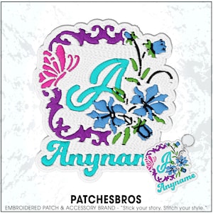 May include: An embroidered patch featuring a large turquoise letter A, a pink butterfly, and blue flowers framed by a purple decorative border. Below the letter, the text Anyname is embroidered in turquoise. A matching keychain is also visible.