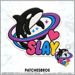 May include: An embroidered patch featuring a black and white orca whale swimming through a purple and blue ringed planet, accompanied by a pink heart and the word SLAY in pink, purple, blue, and yellow letters, with a smaller matching keychain.