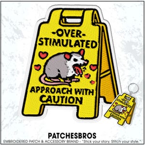 May include: Embroidered yellow caution sign patch with the text "-OVER-STIMULATED APPROACH WITH CAUTION" and a cartoon opossum. The opossum is gray and white with its tongue out. Includes a matching keychain.