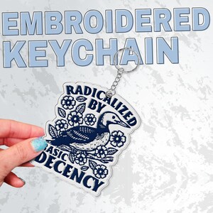 May include: Embroidered keychain with a loon design, surrounded by flowers and leaves, and the text "RADICALIZED BY BASIC DECENCY". The keychain is white with navy blue embroidery and a silver chain.