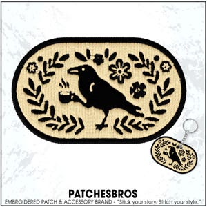 May include: Embroidered patch featuring a black raven holding a cup, surrounded by floral and leaf designs. The patch is oval-shaped with a cream-colored background and black border. A matching keychain is also visible.