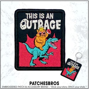 May include: Embroidered patch with the text "THIS IS AN OUTRAGE" above a cartoon image of a hamster wearing a crown riding a blue dinosaur. The patch is black with red and white accents. A matching keychain is also visible.