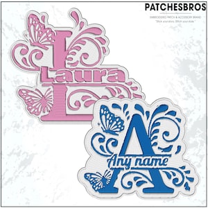 May include: Embroidered patches featuring a butterfly and floral design. One patch is pink with the name "Laura", and the other is blue with the text "Any name". The patches are from the brand "PATCHESBROS".