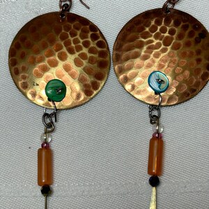 May include: A pair of hammered copper earrings with a round shape and a dangling charm. The charms feature a small green and a blue button, a clear bead, a pink bead, and a brown bead.