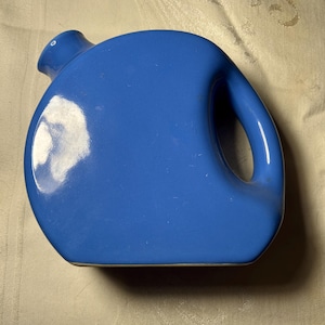 May include: A cobalt blue ceramic pitcher with a rounded body and a large handle. The pitcher has a small spout and a smooth, glossy finish. The design is simple and modern, suitable for serving beverages.