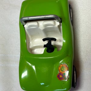 May include: A green toy car with a white interior and a black steering wheel. The car has a silver roll bar and a sticker with flowers on the side.