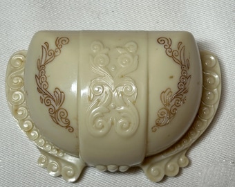 May include: A white plastic ring box with a decorative design in gold. The box is shaped like a half-circle and has a hinged lid.