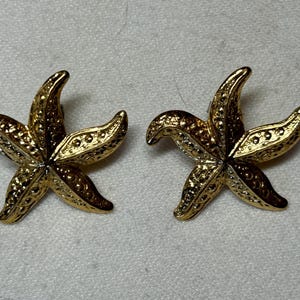May include: A pair of gold tone starfish earrings. The starfish are textured and have a two-tone finish.