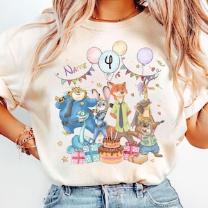 May include: Cream-colored t-shirt featuring a colorful birthday scene with cartoon characters, balloons, and a cake. The number "4" is displayed on a balloon, and the word "Name" is written above the characters. Presents and stars are also included.