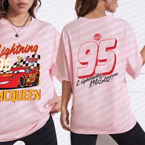 Puede incluir: Camiseta rosa claro con Lightning McQueen de la pel&iacute;cula Cars. El frente muestra el coche, el n&uacute;mero 95 y la frase "Lightning McQueen". La parte trasera tiene un gran "95" rojo y "Lightning McQueen".