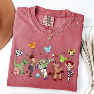 Puede incluir: Una camiseta Comfort Colors de color granate con un gr&aacute;fico colorido de personajes de dibujos animados y globos. El dise&ntilde;o incluye personajes como Woody, Buzz Lightyear y Jessie. Un collar plateado con un colgante est&aacute; sobre la camisa.