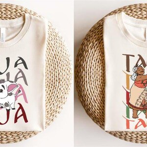 May include: Two cream-colored t-shirts with graphic designs. One features a cartoon pig with the word "PUA" in gray, pink, and maroon. The other shows a woman with the word "TALA" in brown, green, and orange. Both shirts are on a woven surface.