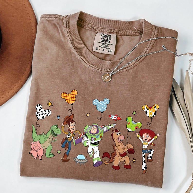 Puede incluir: Una camiseta marr&oacute;n con un gr&aacute;fico colorido de personajes de dibujos animados. El dise&ntilde;o incluye personajes de dibujos animados, globos y estrellas. La camiseta tiene cuello redondo y mangas cortas. La etiqueta dice "Comfort Colors". Tambi&eacute;n se ve un collar plateado.