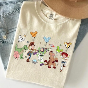 Puede incluir: Camiseta color crema con un gr&aacute;fico colorido de personajes animados sosteniendo globos. El dise&ntilde;o incluye un dinosaurio, un cerdo, un vaquero, un astronauta, un caballo y una vaquera. La camiseta est&aacute; sobre unos vaqueros azules.