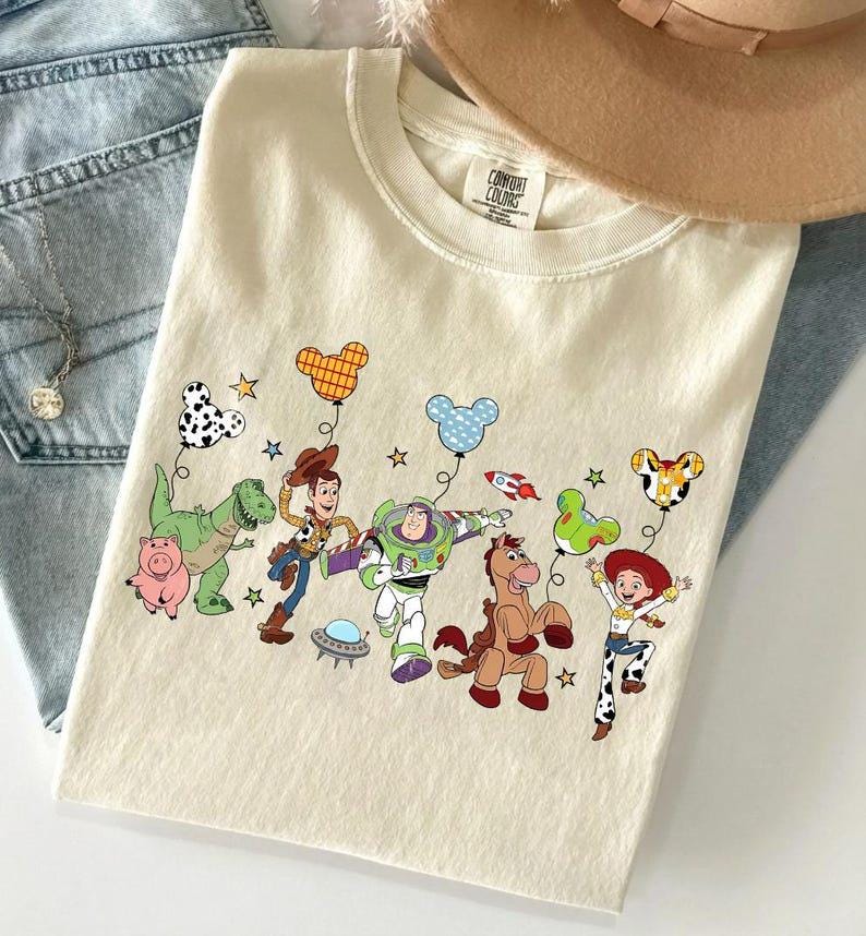 Puede incluir: Camiseta color crema con un gr&aacute;fico colorido de personajes animados sosteniendo globos. El dise&ntilde;o incluye un dinosaurio, un cerdo, un vaquero, un astronauta, un caballo y una vaquera. La camiseta est&aacute; sobre unos vaqueros azules.