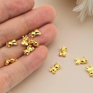 May include: Small, gold-colored bear charms in various poses. The charms are scattered on a light-colored surface, with some held in a hand. The bears have a shiny finish and are detailed with small features.
