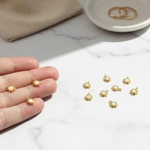 May include: Gold-colored starburst earrings with white centers are displayed on a marble surface. Several earrings are scattered, with a few held in a hand. A small beige pouch and a white dish with gold rings are also visible.