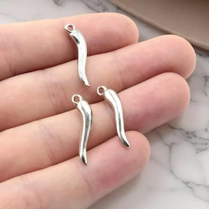 Puede incluir: Tres dijes plateados con forma de pimiento picante, con un lazo en la parte superior para hacer joyas. Los dijes se muestran sobre un fondo neutro, mostrando su forma curva y brillo metálico. Ideales para manualidades.