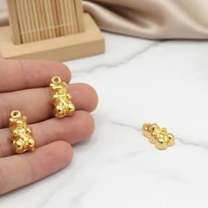May include: Gold-colored gummy bear charms. Three charms are visible, with two held in a hand. The charms have a loop at the top for attaching to jewelry. The background is a white surface with a wooden stand.