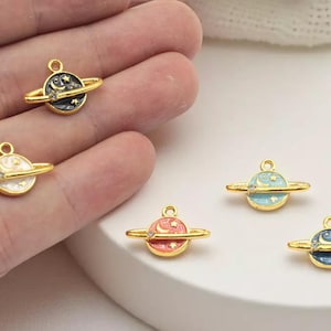 May include: Gold-colored planet charms with colorful enamel details. Each charm features a planet with a ring and a loop for attaching to jewelry. The planets are painted in various colors, including blue, pink, white, and black.