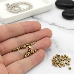 May include: Close-up of small, gold-colored metal beads. Some beads are arranged in a line, while others are in a pile. A silver chain is in a white dish in the background. Black stones are also visible.