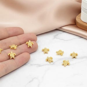 May include: Gold-coloured turtle charms scattered on a white surface and held in a hand. The charms are small, detailed, and have a three-dimensional design. A white jewellery box with a gold zip is in the background.