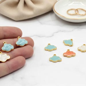 May include: Cloud-shaped charms in various colors, including light blue, white, and coral, with gold-colored edges. Some charms are held in a hand, while others are arranged on a white surface. A small beige pouch and a white dish with gold rings are in the background.