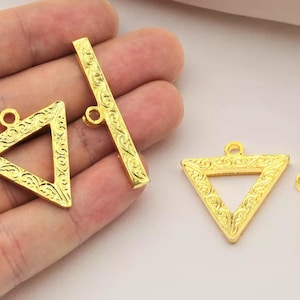 Puede incluir: Componentes de joyería dorados, que incluyen dos colgantes en forma de triángulo y dos cierres en forma de barra. Cada pieza presenta un diseño grabado y ornamentado y un pequeño lazo para la fijación. Los componentes son probablemente para collares o pulseras.