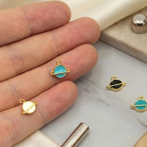 May include: Gold-toned planet charms with colorful enamel details. The charms feature a planet design with a gold band around the center. The planets are in white, black, and turquoise blue. These charms are ideal for jewelry making.