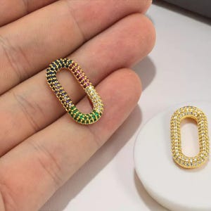 Könnte beinhalten: Zwei goldfarbene, ovale Schmuckanhänger. Ein Anhänger wird in einer Hand gehalten und ist mit farbigen Edelsteinen in einem Regenbogenmuster besetzt. Der andere Anhänger befindet sich auf einer weißen Oberfläche und ist mit klaren Edelsteinen bedeckt.