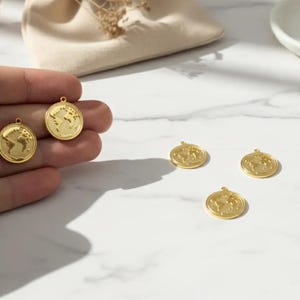 May include: Gold-colored round pendants featuring a detailed world map design. The pendants have a small loop at the top for attaching to a chain or other jewelry. Several pendants are visible, some held in a hand, others scattered on a white surface.