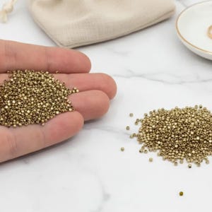 Puede incluir: Un conjunto de pequeñas cuentas de semillas doradas. Algunas cuentas se sostienen en una mano, mientras que otras están dispersas sobre una superficie blanca. También se ve una pequeña bolsa de cordón beige y un plato blanco con dos anillos dorados.