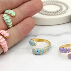 May include: Gold-toned rings with colorful enamel bands in light blue, pink, and lavender. The rings are open-ended and wrap around the finger. Some rings are on a hand, while others are displayed on a white surface.