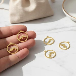 May include: Gold-colored mountain charm pendants. The pendants feature a mountain range silhouette within a circular frame. Several pendants are displayed on a white surface, with a small beige pouch and a white dish in the background.