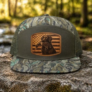 May include: A camouflage-patterned trucker hat with a brown leather patch featuring a dog holding a duck against an American flag design. The hat has a green mesh back and a curved brim.
