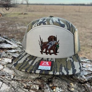May include: A camouflage flat bill snapback hat with a brown dog graphic carrying a duck. The hat has a tan front panel and a mesh back. The brim and the rest of the hat have a camouflage pattern. The hat has a tag attached.