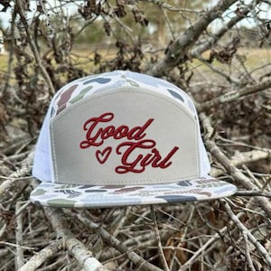 May include: A camouflage print trucker hat with a white mesh back and a flat brim. The front panel is tan with the words "Good Girl" embroidered in red script with a heart symbol.