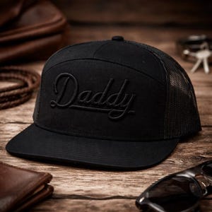 May include: Black trucker hat with the word "Daddy" embroidered in a script font. The hat has a mesh back and a flat brim. The hat is sitting on a wooden surface.