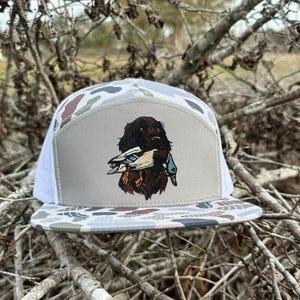 May include: A camouflage-patterned trucker hat with a white mesh back and a flat brim. The front panel features a brown dog with a duck in its mouth. The hat's design incorporates shades of gray, brown, and green.