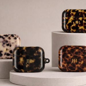 May include: Four AirPod Pro cases with a tortoiseshell pattern in shades of brown, black, and cream. Each case has gold-colored initials "JH" and a small loop. The cases are displayed on white and gray pedestals.