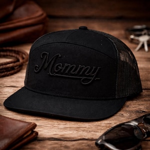 May include: Black trucker hat with the word "Mommy" embroidered in a cursive font on the front. The hat has a mesh back and a curved brim. The hat is sitting on a wooden surface.